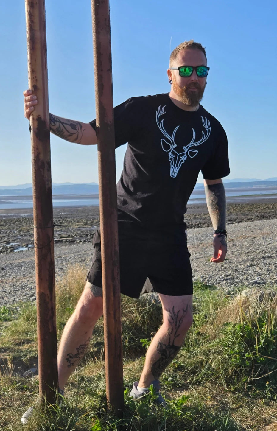 Man standing outdoors by a beach holding two wooden poles, wearing a black t-shirt with a deer design.