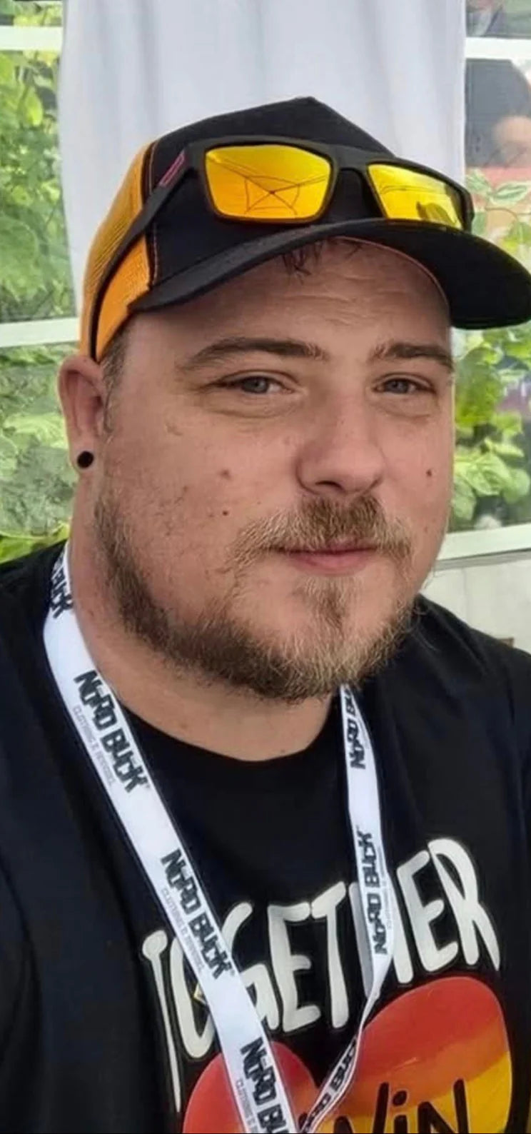 Michael the owner wearing a cap with sunglasses on top, black t-shirt with visible text, and lanyard.