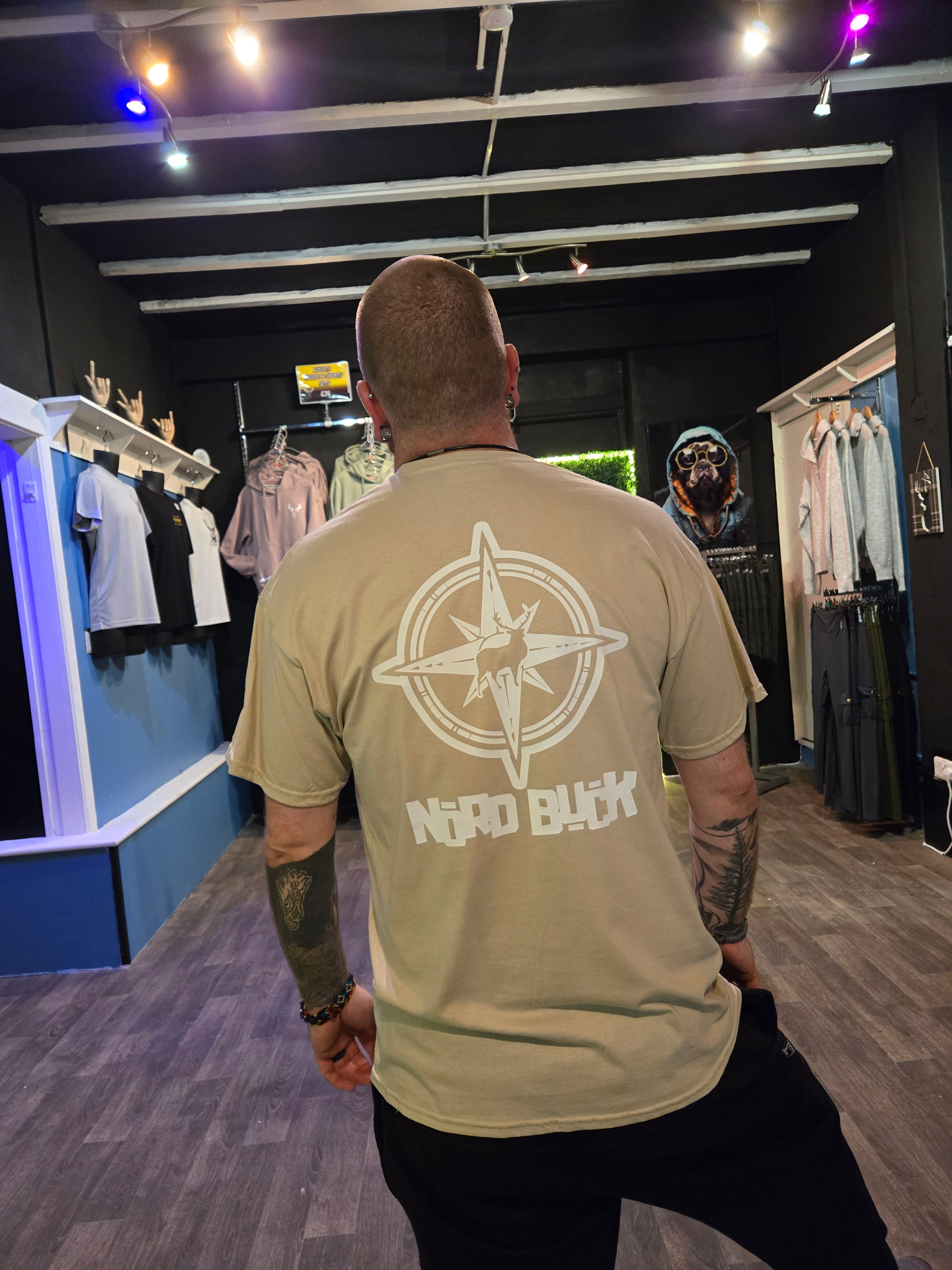Person wearing a beige t-shirt with a compass logo and text, standing in an indoor setting.