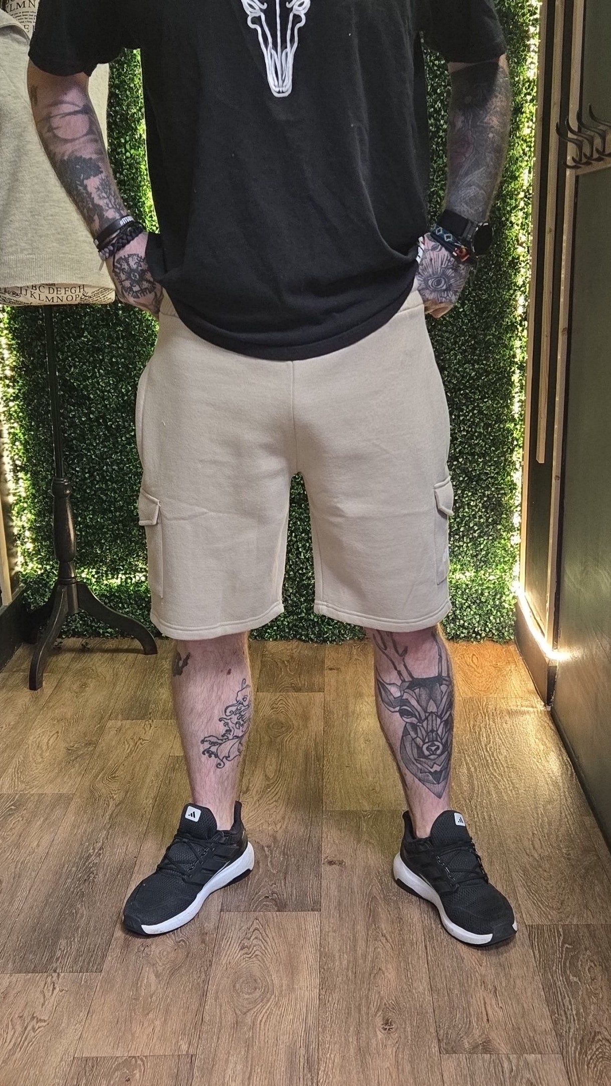 Person wearing a black t-shirt, beige cargo shorts, and black sneakers standing indoors.