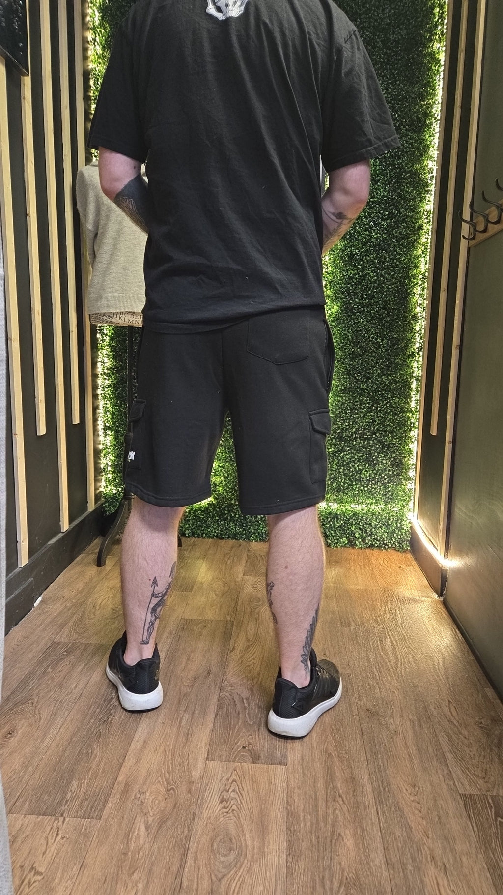 Person wearing black shorts and a black shirt standing indoors with greenery in the background.
