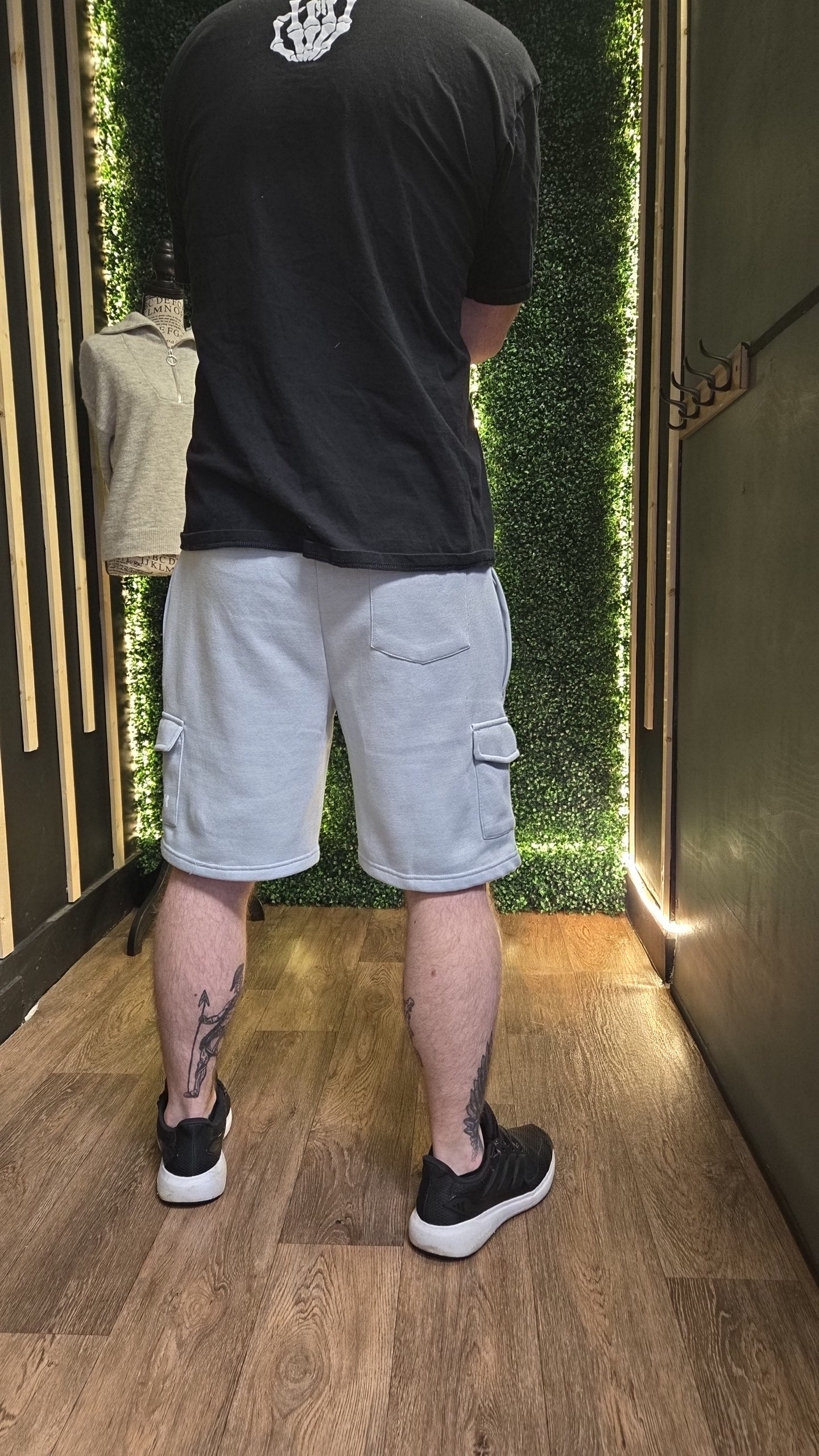Person wearing a black t-shirt and light gray shorts standing in a room with greenery.