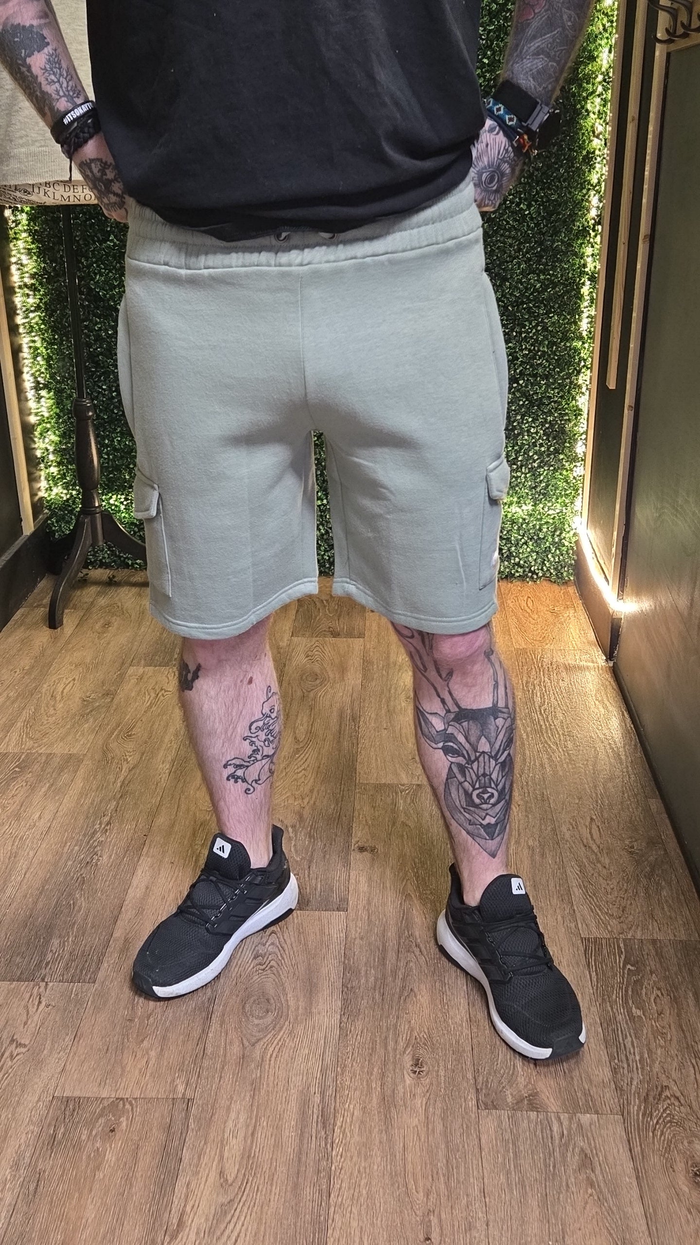 Person wearing light gray shorts and black sneakers on a wooden floor with greenery in the background