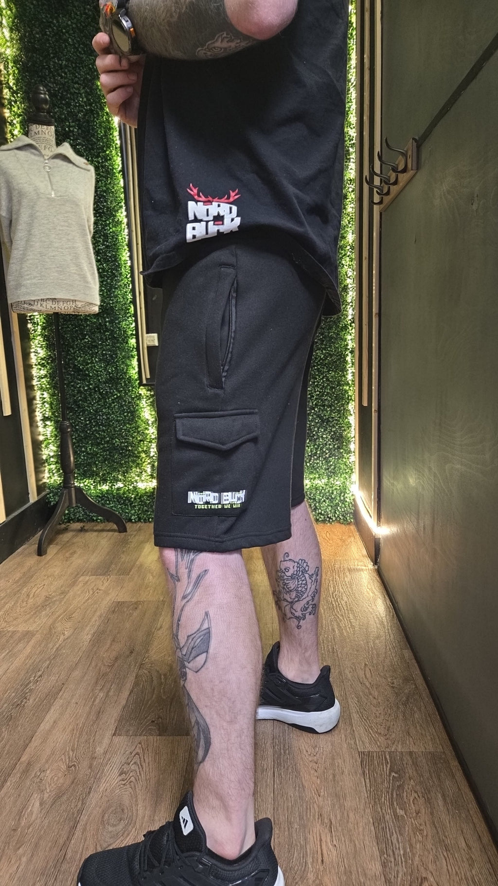 Person wearing black shorts with visible branding in a room with greenery and wooden floor.