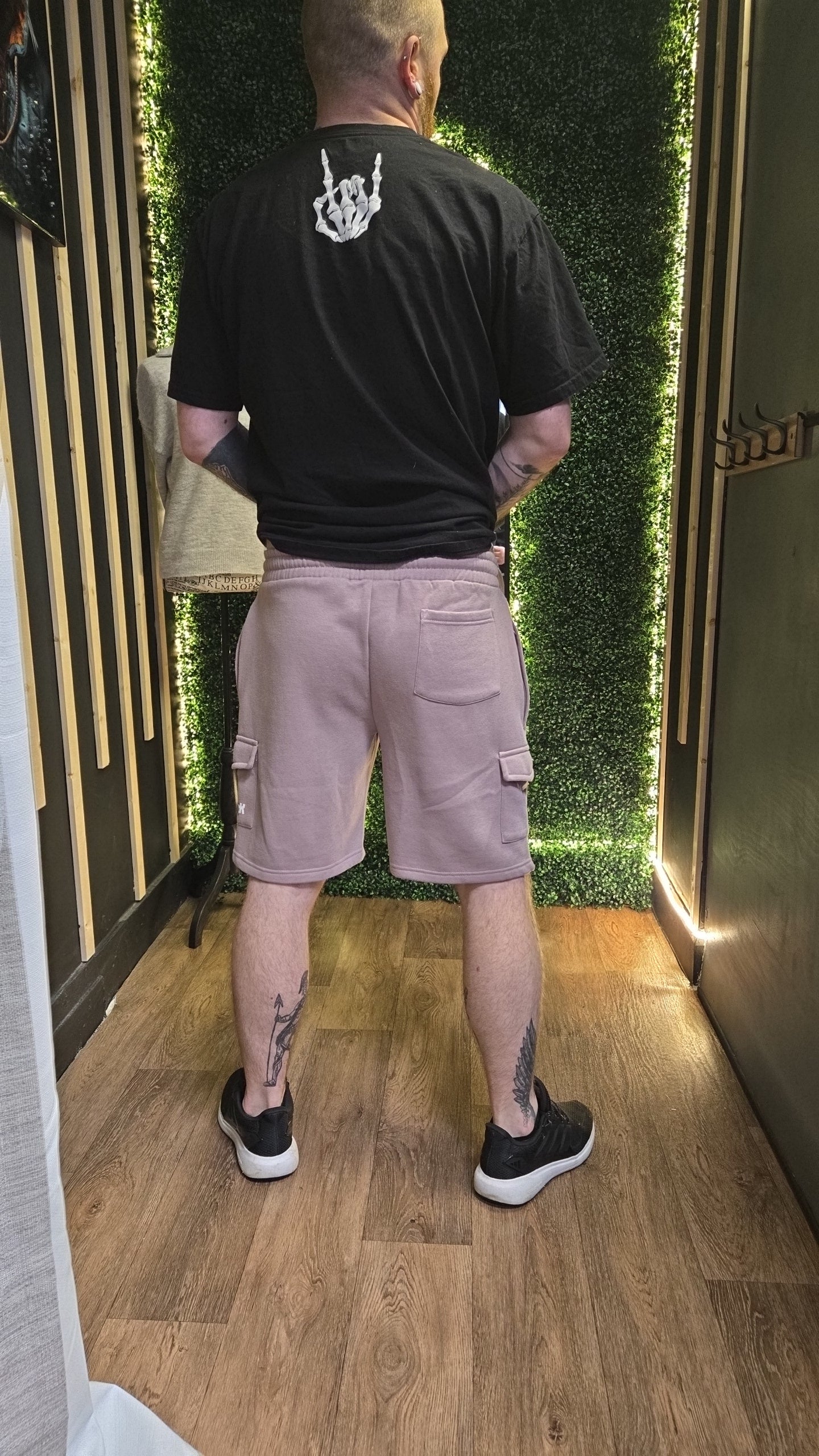 Person wearing a black t-shirt and pink shorts standing in a room with wooden flooring and green wall.