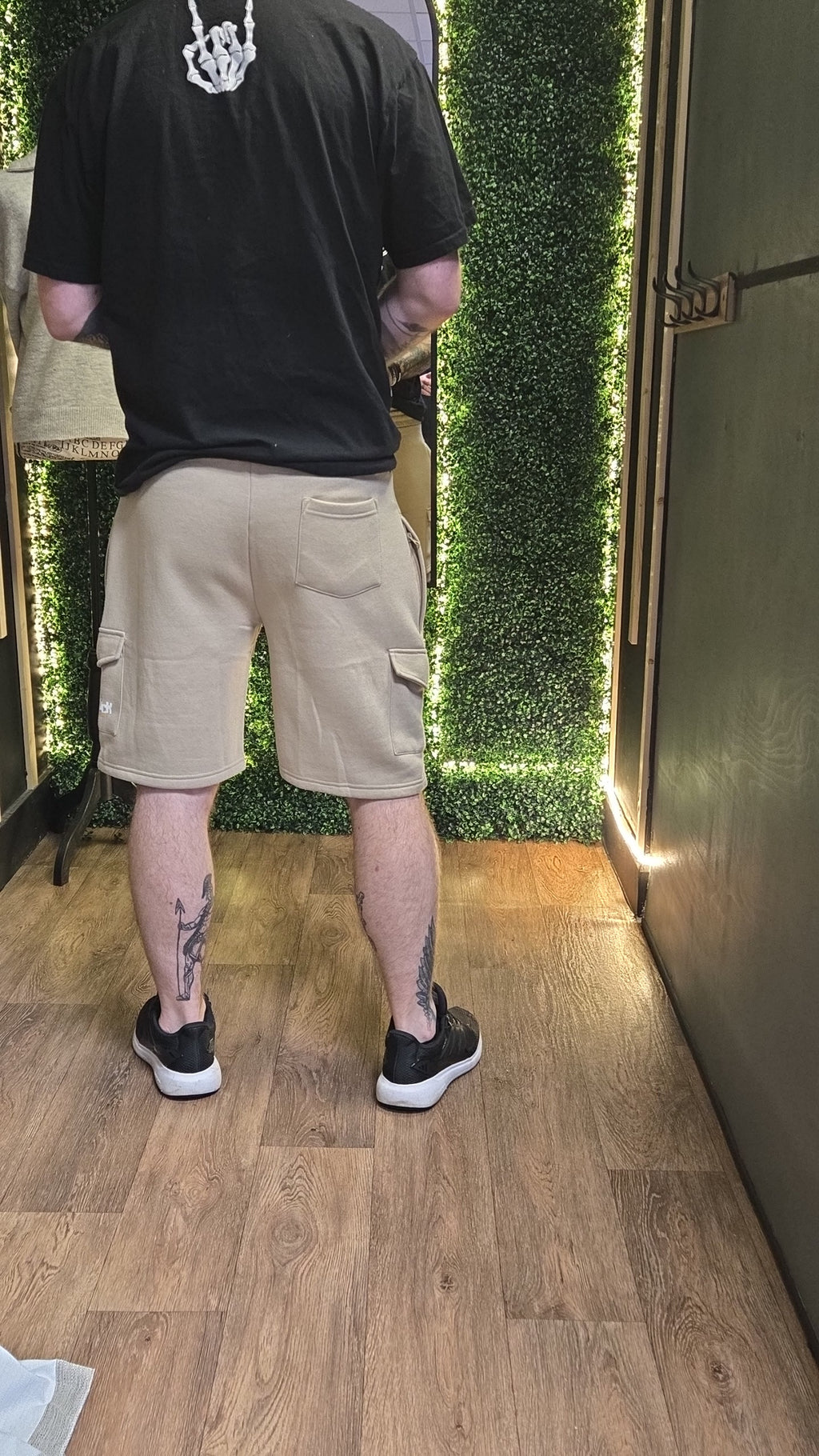 Person wearing a black t-shirt and beige cargo shorts standing in a room with greenery.
