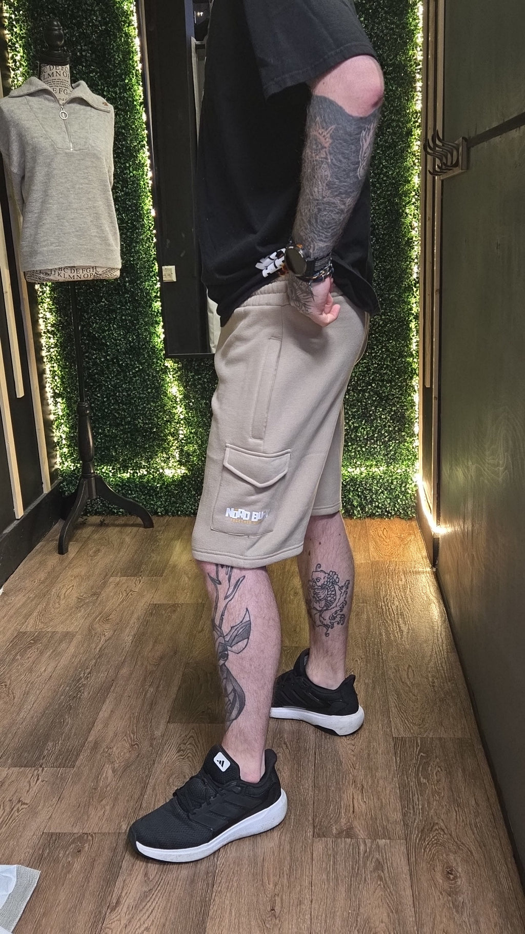 Person wearing beige cargo shorts and black sneakers with tattoos on a wooden floor.