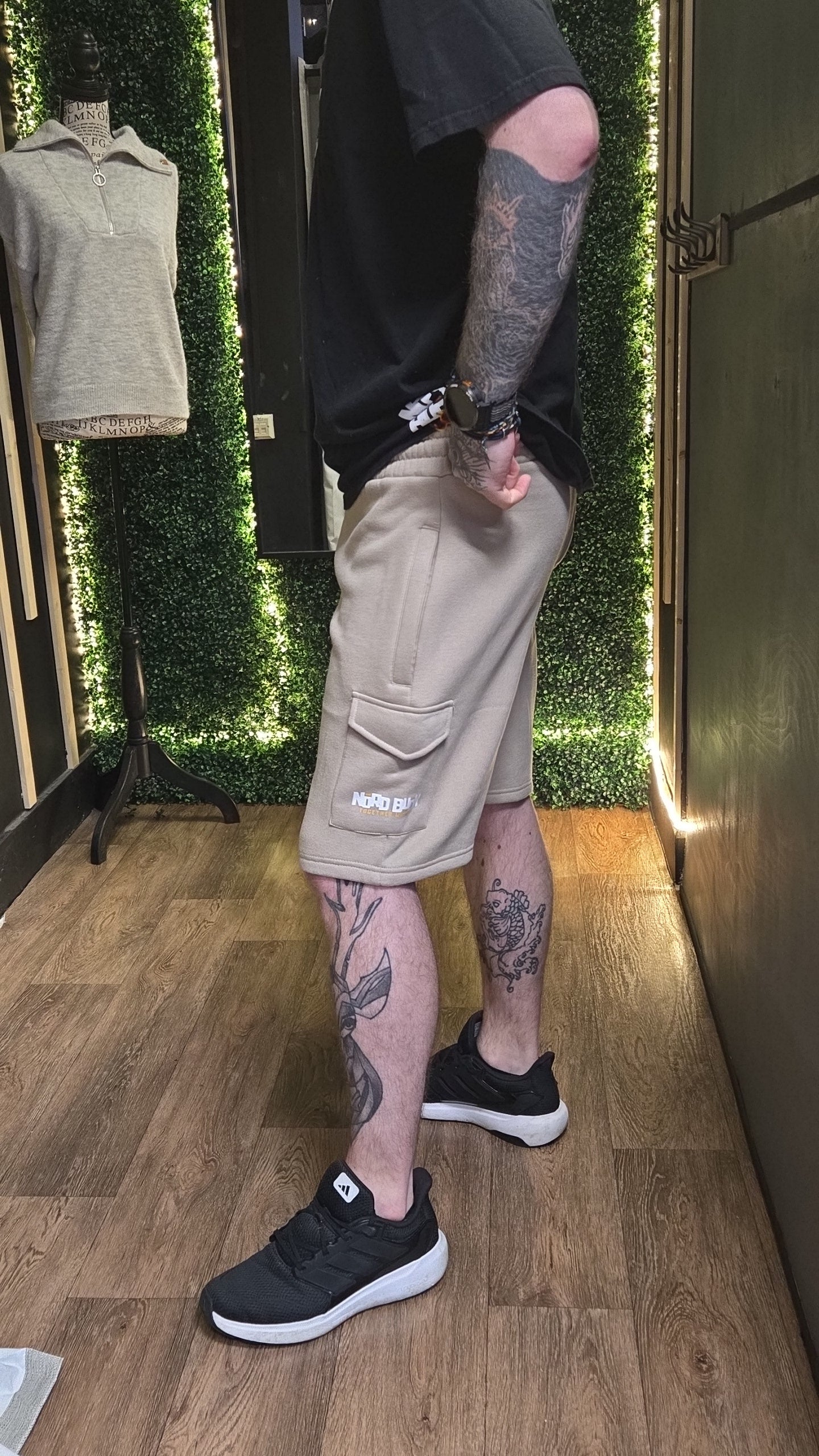 Person wearing beige cargo shorts and black sneakers with tattoos on a wooden floor.