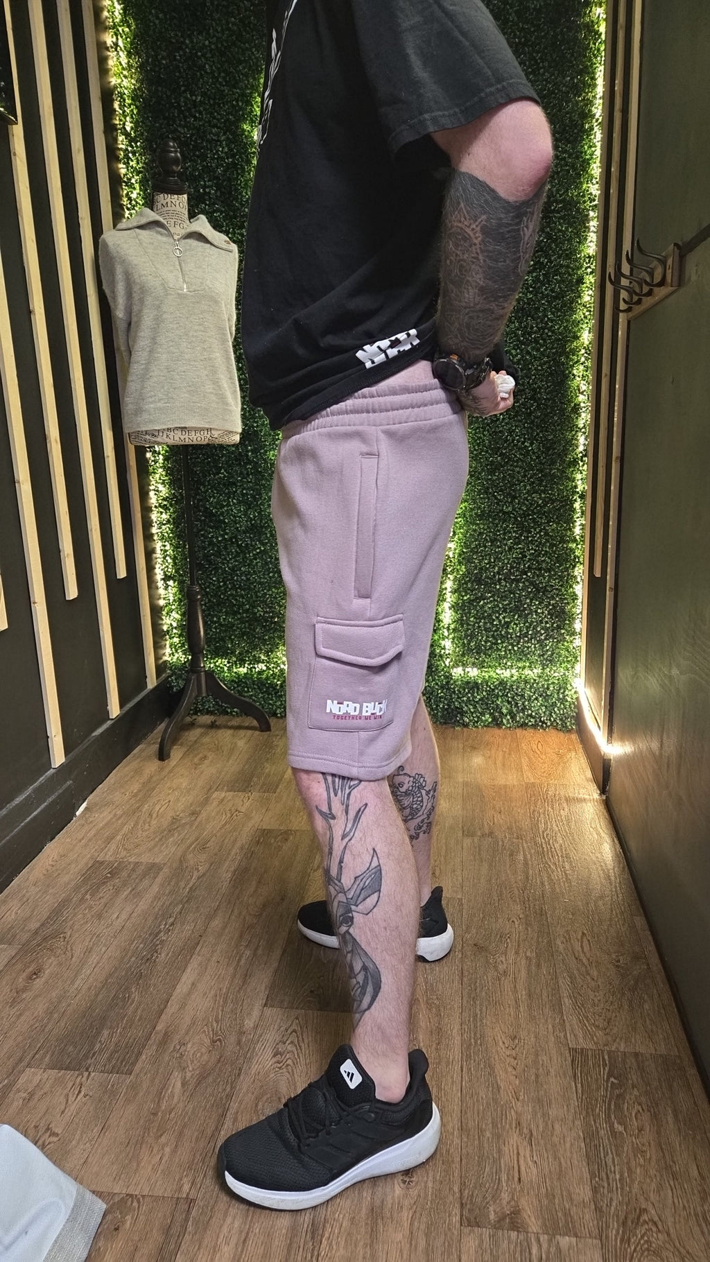 Person wearing a black t-shirt, light Lavender shorts, and black sneakers with tattoos on arms and legs in a store setting.