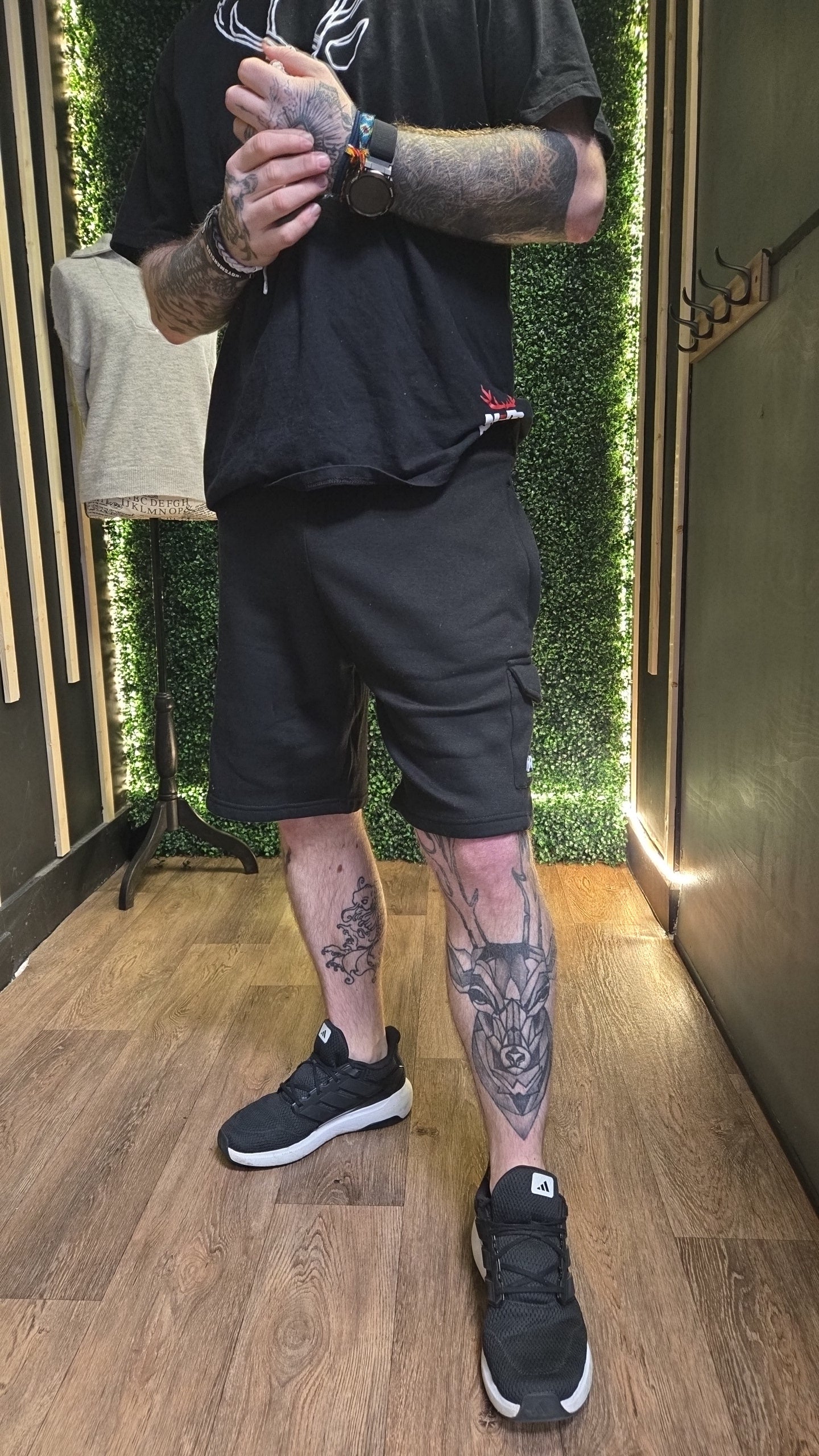 Person wearing black t-shirt, shorts, and sneakers with tattoos on arms and legs in a room with greenery.