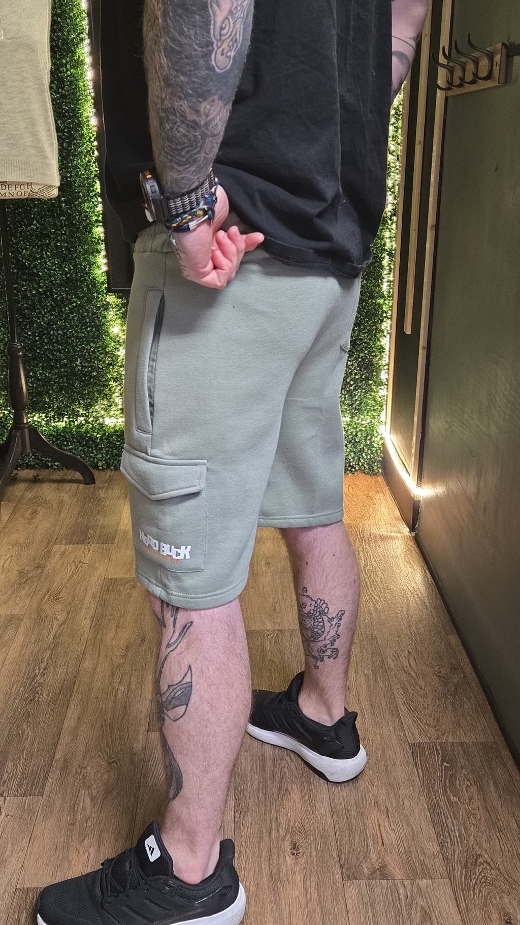 Person wearing light gray cargo shorts with visible tattoos on arms and legs, standing indoors.