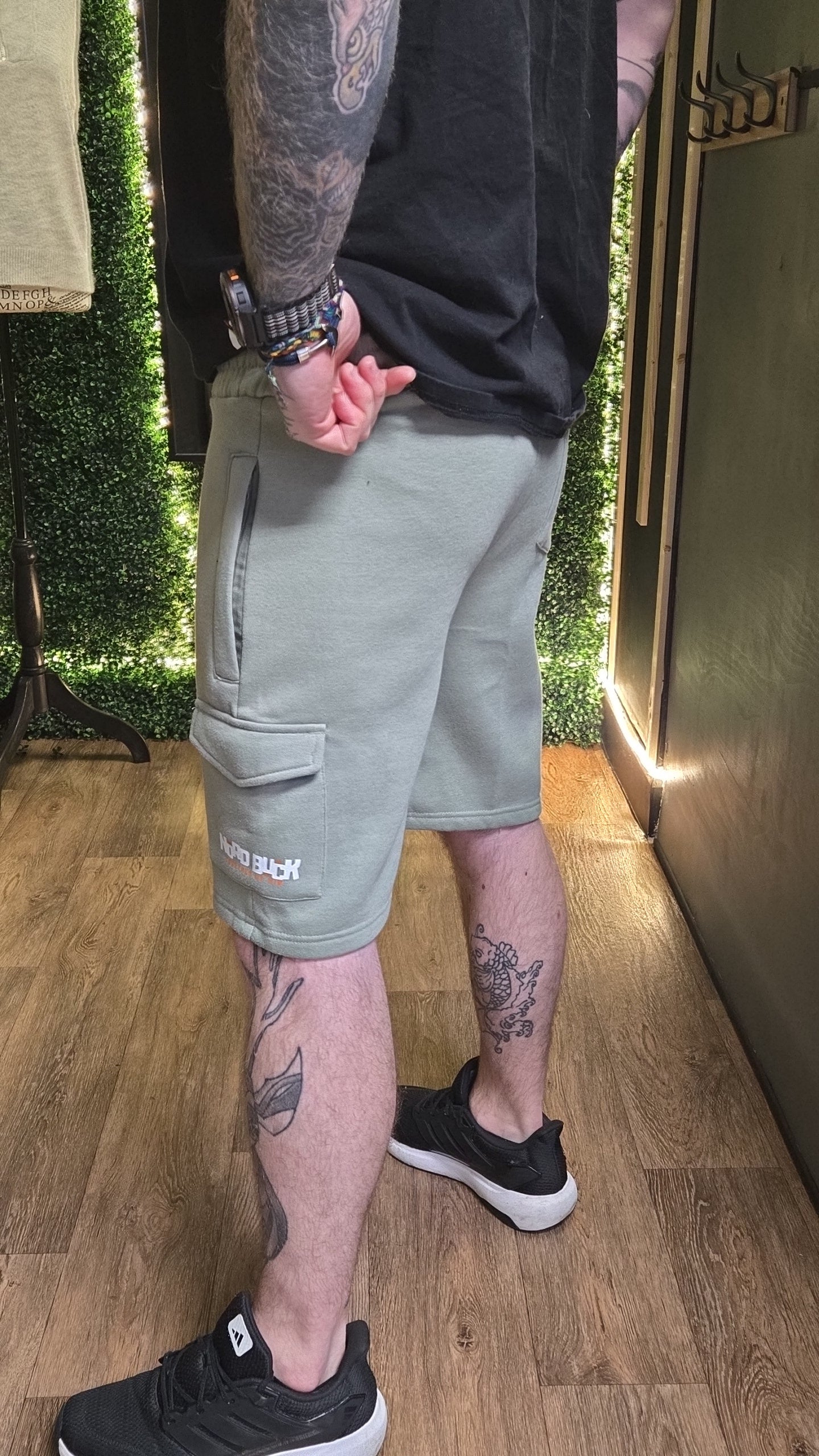 Person wearing light gray cargo shorts with visible tattoos on arms and legs, standing indoors.