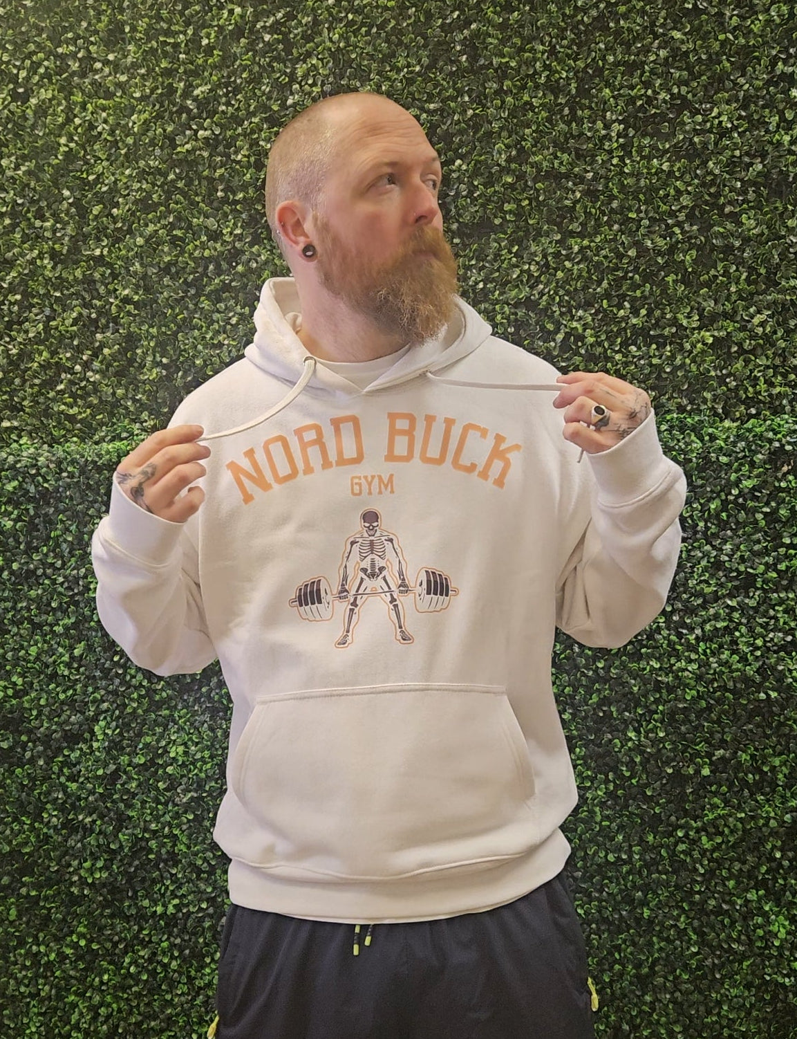 Man wearing a hoodie with 'Nord Buck Gym' design against a green hedge background