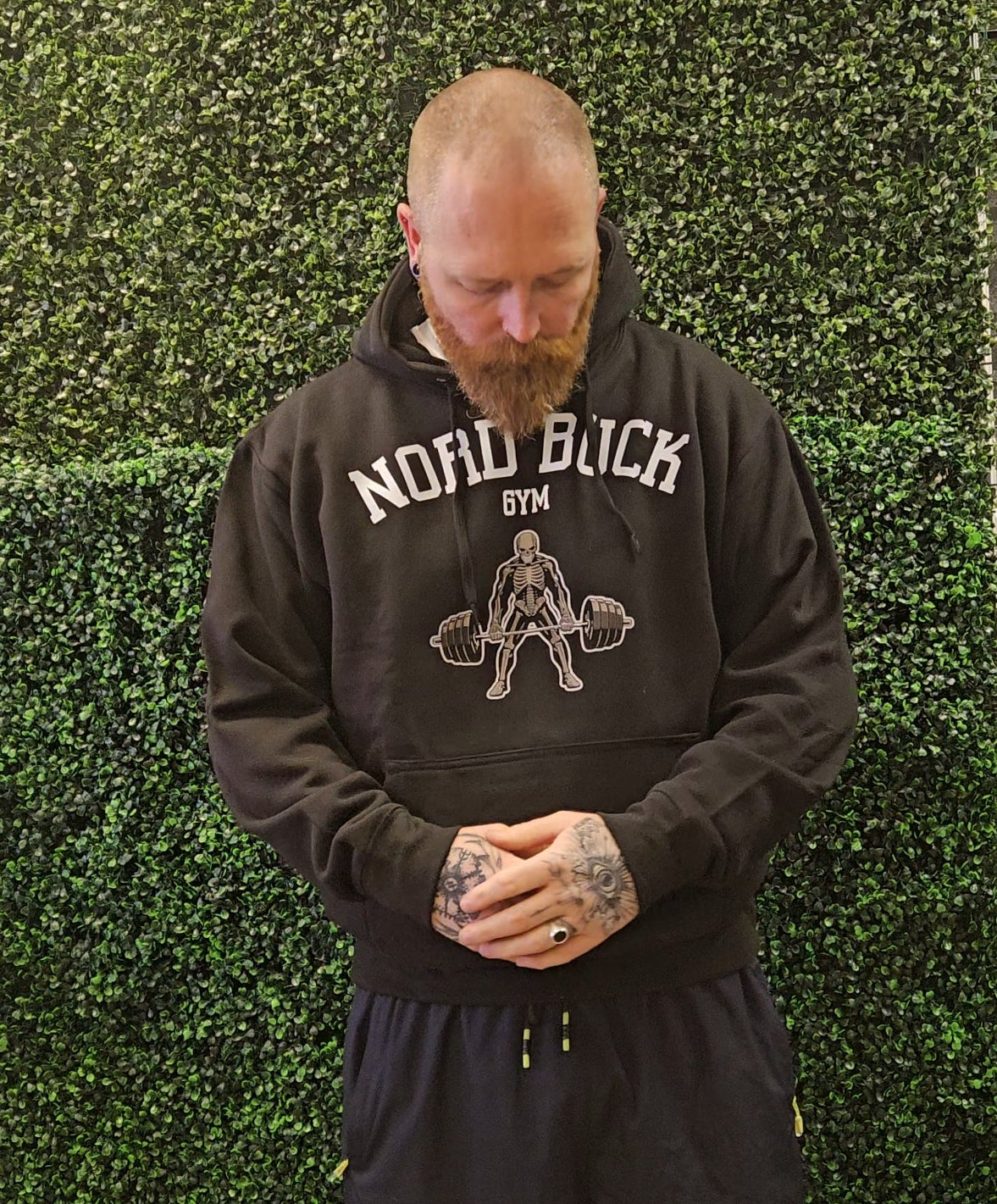 Man wearing a black hoodie with 'Nord Buck Gym' logo against a green textured wall.