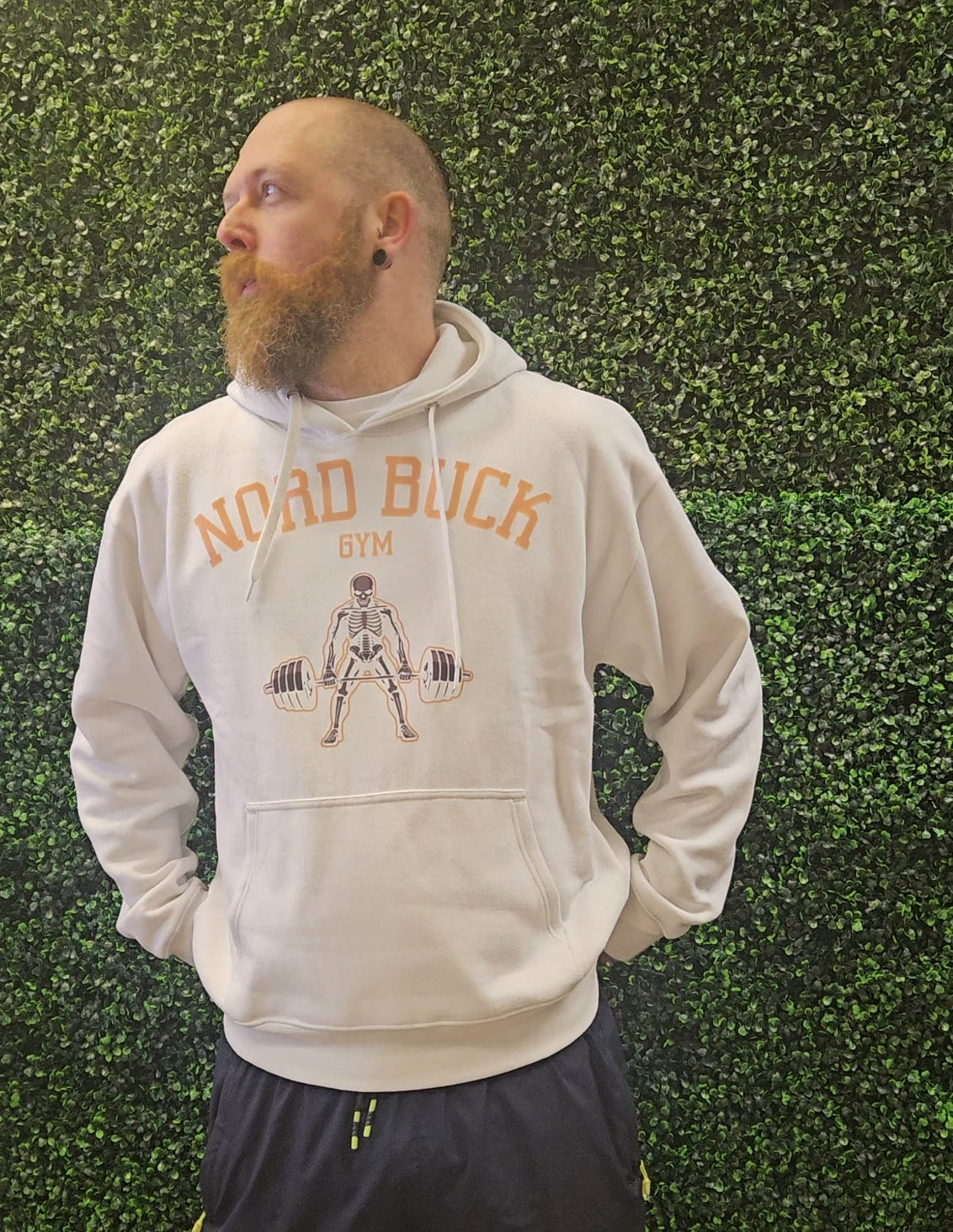 Man wearing a hoodie with 'Nad Buck Gym' design against a green hedge background